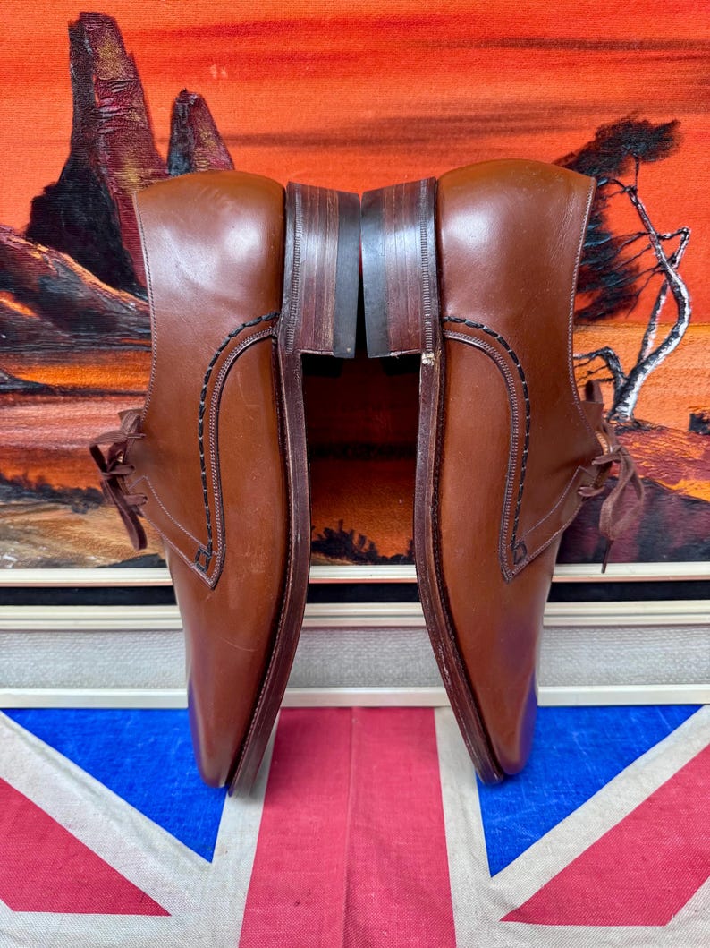May include: A pair of brown leather dress shoes with laces. The shoes have a classic design with stitched detailing and a polished finish. The background includes a painting with orange and red tones and a Union Jack flag.