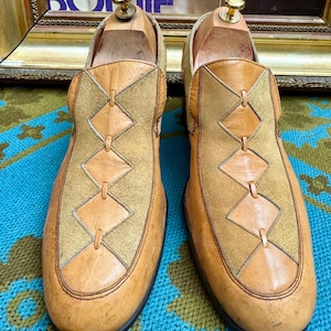 May include: A pair of tan leather slip-on shoes with a geometric design on the vamp. The shoes feature a light brown suede panel with diamond-shaped leather accents. The soles are black, and the shoes are on wooden shoe trees.