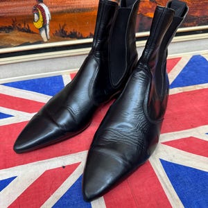 May include: A pair of black leather Chelsea boots with pointed toes, smooth uppers, and black elastic side panels. The boots feature a high-cut ankle design with a pull tab at the back of each heel and a sleek, polished finish on the leather surface.