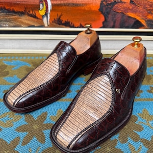 Op de afbeelding: Een paar bruine leren instappers met een reptielachtige textuur en een lichtbeige middenpaneel. De schoenen hebben een vierkante neus en worden gepresenteerd op een blauwgroen oppervlak met een patroon. Houten schoenspanners zitten erin.