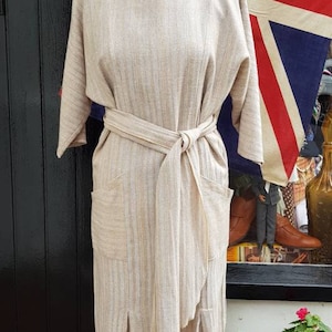 Puede incluir: Un vestido de lino beige con estampado de rayas, cinturón de anudar y dos bolsillos. El vestido tiene un corte holgado y una longitud midi.
