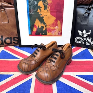 May include: A pair of brown leather shoes with black laces, set on a Union Jack flag. The shoes have a unique design with vertical and horizontal stitched accents. A framed artwork and two Adidas bags are in the background.