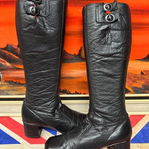 Puede incluir: Un par de botas negras hasta la rodilla con puntera cuadrada y tacón ancho. Cada bota tiene dos hebillas plateadas en la parte superior. Las botas son de cuero texturizado y tienen una estética vintage.