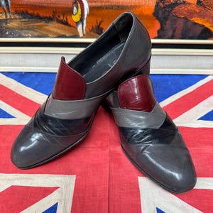 K&ouml;nnte beinhalten: Ein Paar graue Lederschuhe mit geometrischem Design. Die Schuhe haben einen weinroten Akzent am Blatt und ein schwarz-graues Muster &uuml;ber der Zehenpartie. Die Schuhe werden auf einer Union Jack-Flagge pr&auml;sentiert.