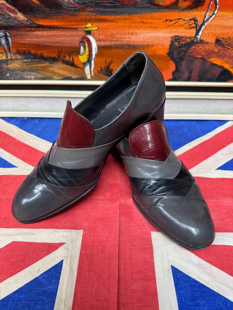 K&ouml;nnte beinhalten: Ein Paar graue Lederschuhe mit geometrischem Design. Die Schuhe haben einen weinroten Akzent am Blatt und ein schwarz-graues Muster &uuml;ber der Zehenpartie. Die Schuhe werden auf einer Union Jack-Flagge pr&auml;sentiert.