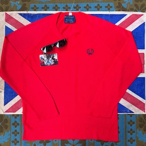 May include: A bright red long-sleeved sweater with a crew neck, featuring a small navy blue emblem on the chest. A pair of black sunglasses and a small photograph are placed on the sweater. The sweater is displayed on a Union Jack flag.