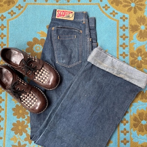 Puede incluir: Un par de pantalones vaqueros de mezclilla azul oscuro con una etiqueta roja y blanca que dice "SCORPION" en el bolsillo trasero. Los pantalones están doblados en la parte inferior y están sobre una alfombra de patrón azul y dorado. Un par de zapatos marrones a cuadros están sobre la alfombra junto a los pantalones.