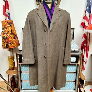 Puede incluir: Un abrigo de lana marrón con un diseño clásico, con solapas y tres botones. Una bufanda azul y morada está drapeada alrededor del cuello. El abrigo se exhibe en un soporte de madera.