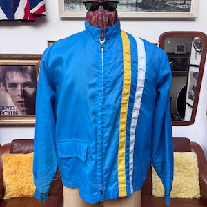 May include: A blue zip-up jacket with a yellow and white stripe down the front. The jacket has two front pockets and a high collar. The jacket is displayed on a mannequin. The jacket is a vintage style.