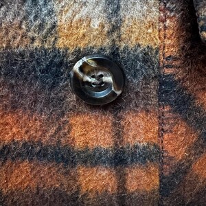 May include: Close-up of a brown, black, and orange plaid fabric with a large, round, marbled button. The button is centered on the image, and the fabric has a textured, woolly appearance.