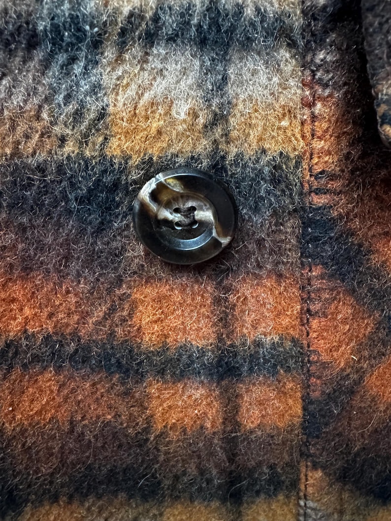 May include: Close-up of a brown, black, and orange plaid fabric with a large, round, marbled button. The button is centered on the image, and the fabric has a textured, woolly appearance.