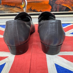 K&ouml;nnte beinhalten: Ein Paar dunkelgraue Leder-Loafer mit kleinem Absatz. Die Schuhe werden auf einer rot-wei&szlig;-blauen Union-Jack-Flagge pr&auml;sentiert. Im Hintergrund befindet sich ein &Ouml;lgem&auml;lde.