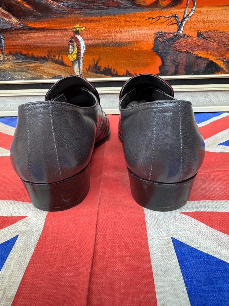 K&ouml;nnte beinhalten: Ein Paar dunkelgraue Leder-Loafer mit kleinem Absatz. Die Schuhe werden auf einer rot-wei&szlig;-blauen Union-Jack-Flagge pr&auml;sentiert. Im Hintergrund befindet sich ein &Ouml;lgem&auml;lde.