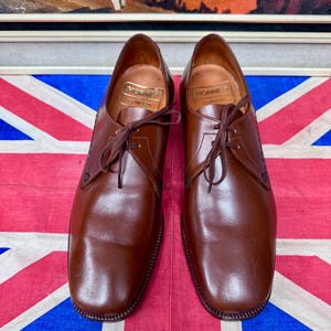 May include: A pair of brown leather dress shoes with laces, displayed on a Union Jack flag. The shoes have a classic design with a rounded toe and a stitched sole. The brand "Hornes London" is visible inside the shoe.