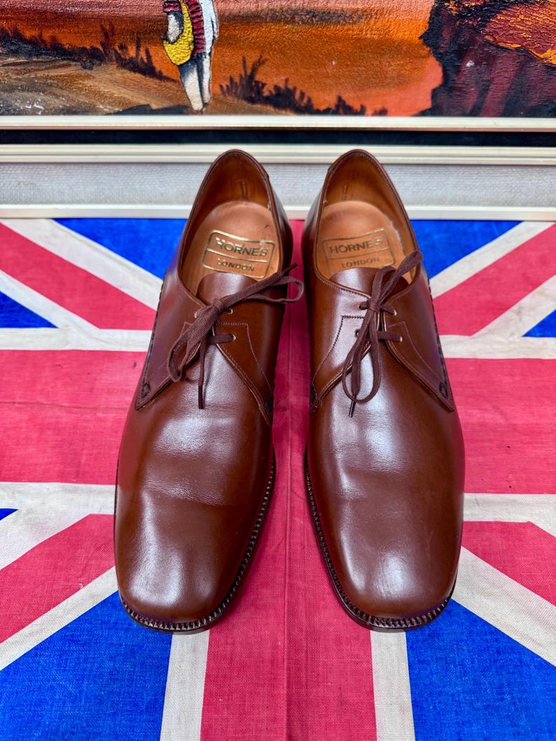 May include: A pair of brown leather dress shoes with laces, displayed on a Union Jack flag. The shoes have a classic design with a rounded toe and a stitched sole. The brand "Hornes London" is visible inside the shoe.