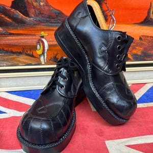 May include: A pair of black leather platform shoes featuring a rounded toe, thick rubber soles, and black lace-up closures. The shoes have decorative stitching patterns on the upper panels and include wooden shoe trees inside the heel area.