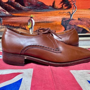 May include: A pair of brown leather dress shoes with a low heel and stitched detailing. The shoes are laced up and sit on a red and blue Union Jack flag. The background features a framed painting.