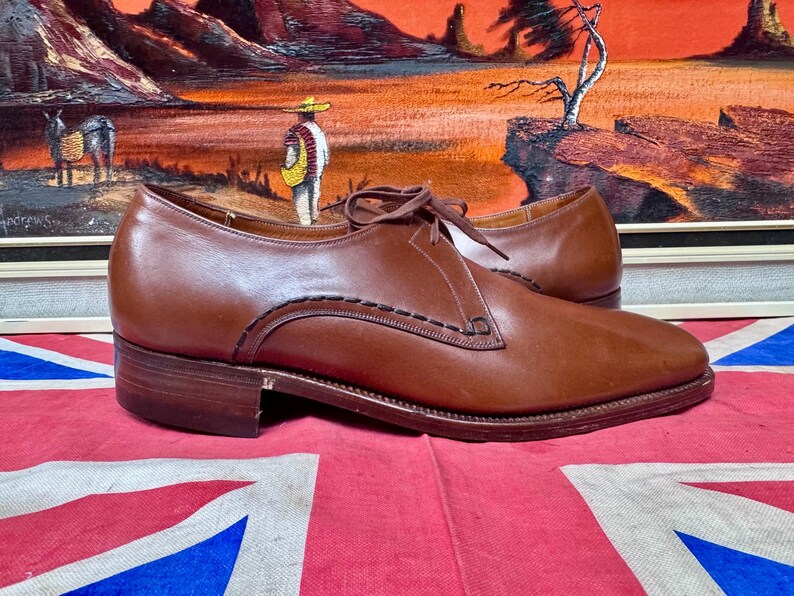 May include: A pair of brown leather dress shoes with a low heel and stitched detailing. The shoes are laced up and sit on a red and blue Union Jack flag. The background features a framed painting.