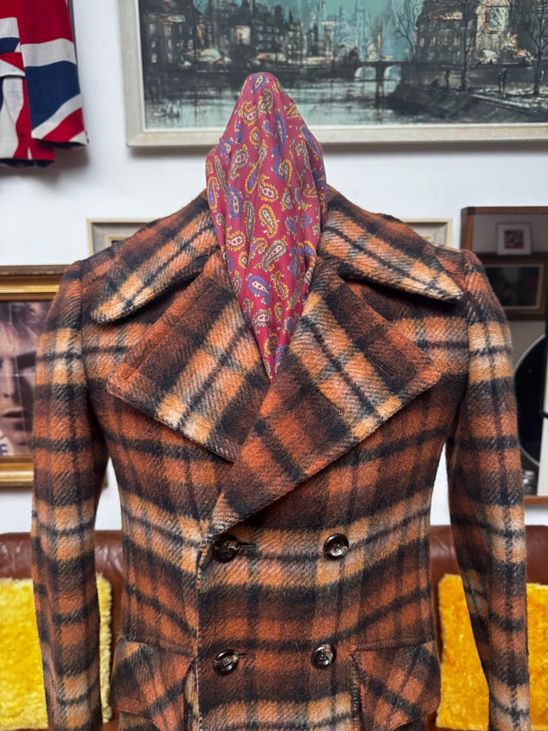 May include: A vintage double-breasted plaid coat in shades of orange, brown, and black. The coat features a classic notched collar and dark buttons. A paisley scarf in red, blue, and yellow is draped over the coat's shoulders.