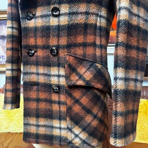 May include: A double-breasted jacket featuring a brown, black, and cream plaid pattern. The garment includes a wide lapel, long sleeves, and a flap pocket on the side. Five dark, round buttons are visible on the front closure of the jacket.