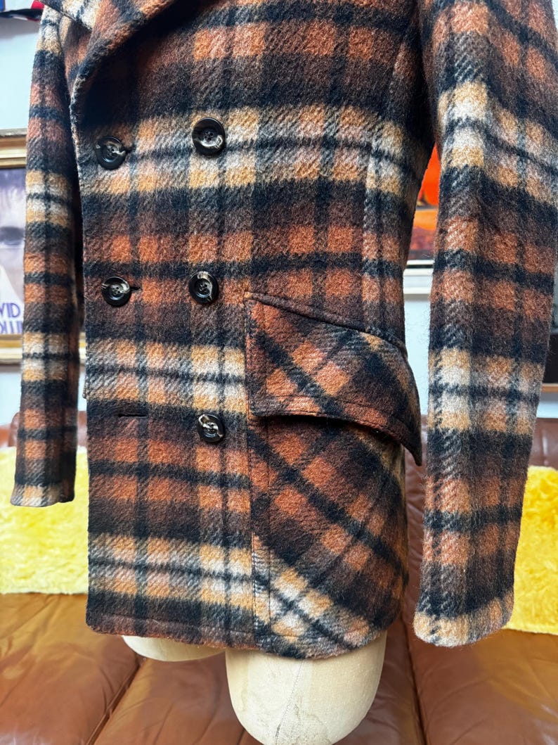 May include: A double-breasted jacket featuring a brown, black, and cream plaid pattern. The garment includes a wide lapel, long sleeves, and a flap pocket on the side. Five dark, round buttons are visible on the front closure of the jacket.