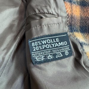 May include: Close-up of the interior of a coat, featuring a brown lining and a label with text. The label reads "80% WOLLE 20% POLYAMID FUTTERSTOFF: 100% VISCOSE" with care symbols. The coat's exterior has a patterned design in orange, brown, and blue.