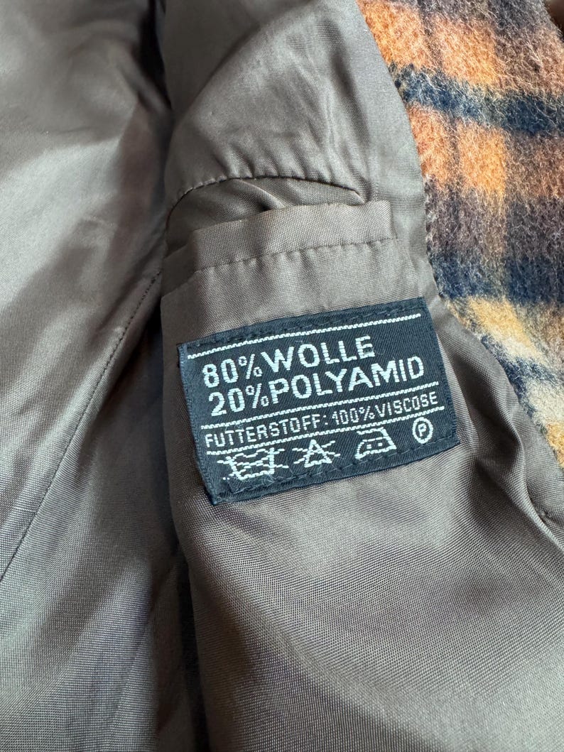 May include: Close-up of the interior of a coat, featuring a brown lining and a label with text. The label reads "80% WOLLE 20% POLYAMID FUTTERSTOFF: 100% VISCOSE" with care symbols. The coat's exterior has a patterned design in orange, brown, and blue.