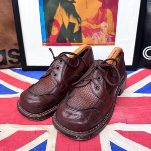 May include: A pair of brown leather shoes with a textured design on the toe and vamp. The shoes have a lace-up closure and a thick sole. The shoes are displayed on a Union Jack flag.