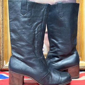 May include: A pair of black leather boots with a mid-height wooden heel. The boots have a tall shaft and a small strap detail at the top. The boots are displayed on a Union Jack flag.