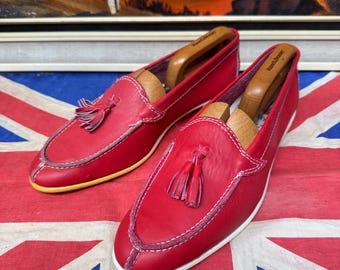 Pode incluir: Um par de mocassins de couro vermelho com costuras brancas e detalhes de borlas. Os sapatos têm bico arredondado e uma borda de sola amarela. Formas de sapato de madeira dentro. Sobre uma bandeira da Union Jack.