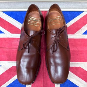 May include: A pair of brown leather dress shoes with laces, displayed on a Union Jack flag. The shoes have a classic design with a rounded toe and the brand name "Hornes London" visible inside. The leather has a polished finish.