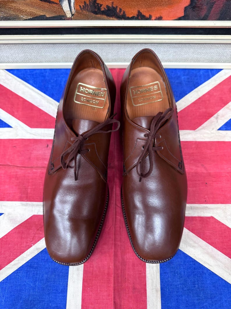 May include: A pair of brown leather dress shoes with laces, displayed on a Union Jack flag. The shoes have a classic design with a rounded toe and the brand name "Hornes London" visible inside. The leather has a polished finish.