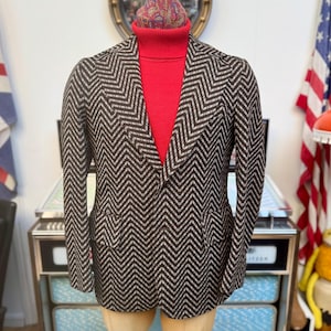 May include: A brown and white herringbone patterned blazer with a red turtleneck sweater underneath. The blazer has a single button closure and a pointed collar.