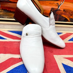 May include: A pair of white leather loafers featuring a block heel with a dark brown wood-tone finish. Each shoe has a small gold-tone metal accent on the vamp. One shoe is positioned upright while the other rests on its side with a wooden shoe tree.