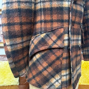 May include: A close-up of a plaid jacket in shades of brown, orange, and black. The jacket has a classic design with a notched lapel, a pocket, and buttons. The fabric appears to be a wool blend.