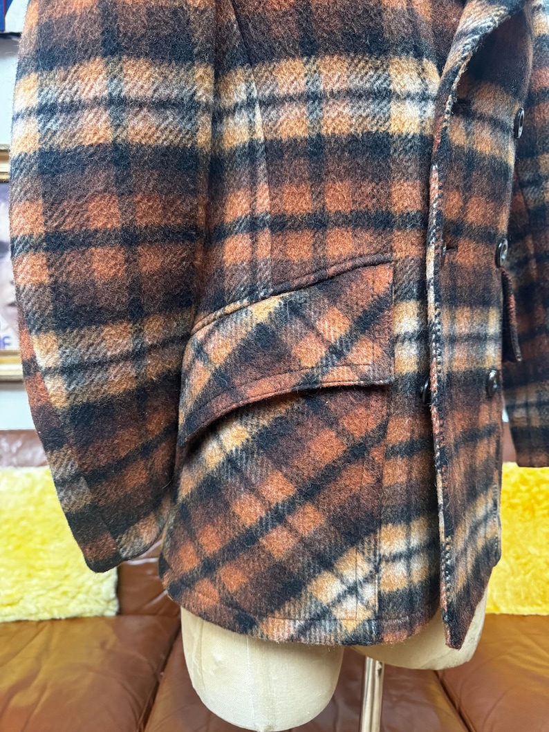 May include: A close-up of a plaid jacket in shades of brown, orange, and black. The jacket has a classic design with a notched lapel, a pocket, and buttons. The fabric appears to be a wool blend.