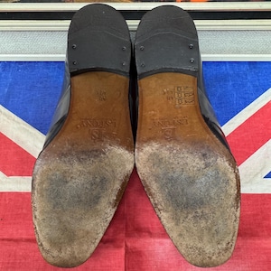 K&ouml;nnte beinhalten: Ein Paar schwarze Lederschuhe, Sohlen nach oben, mit Gebrauchsspuren und dem Markennamen "L.S. FONY". Die Schuhe stehen vor dem Hintergrund einer Union Jack Flagge und einem gerahmten Gem&auml;lde.