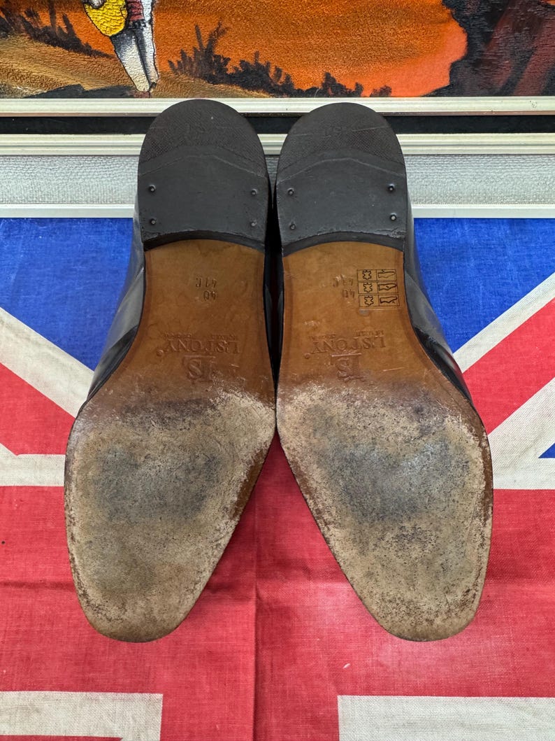 K&ouml;nnte beinhalten: Ein Paar schwarze Lederschuhe, Sohlen nach oben, mit Gebrauchsspuren und dem Markennamen "L.S. FONY". Die Schuhe stehen vor dem Hintergrund einer Union Jack Flagge und einem gerahmten Gem&auml;lde.
