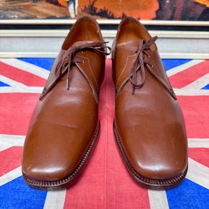 May include: A pair of brown leather dress shoes with laces. The shoes have a classic design with a rounded toe and a stitched sole. The shoes are displayed on a Union Jack flag.