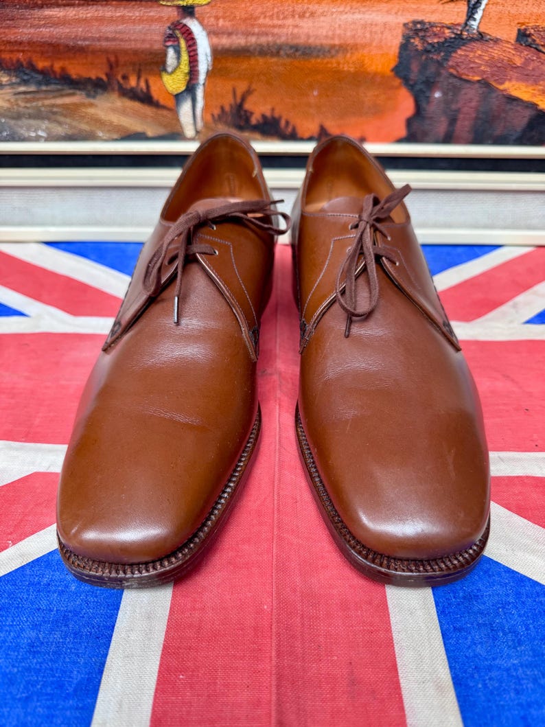 May include: A pair of brown leather dress shoes with laces. The shoes have a classic design with a rounded toe and a stitched sole. The shoes are displayed on a Union Jack flag.