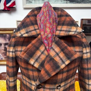 May include: A double-breasted plaid coat in shades of orange and black, featuring a wide collar and button details. A paisley patterned scarf in red, blue, and yellow is tucked into the coat's neckline. The coat is displayed on a brown leather couch.
