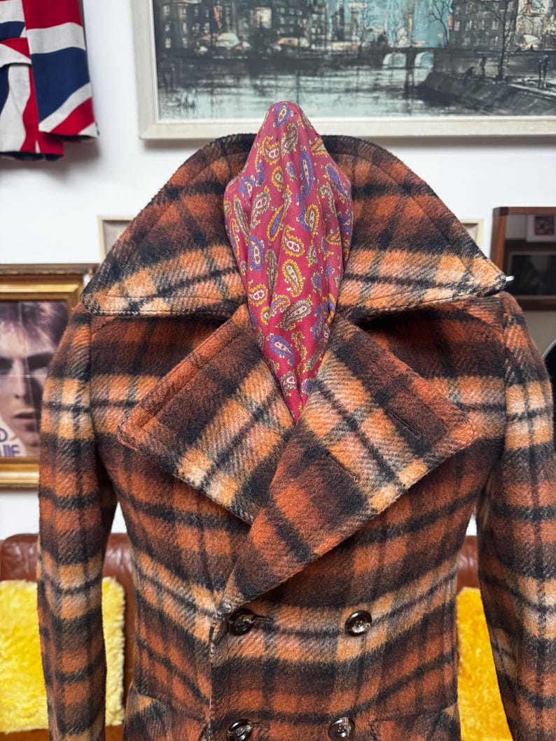May include: A double-breasted plaid coat in shades of orange and black, featuring a wide collar and button details. A paisley patterned scarf in red, blue, and yellow is tucked into the coat's neckline. The coat is displayed on a brown leather couch.