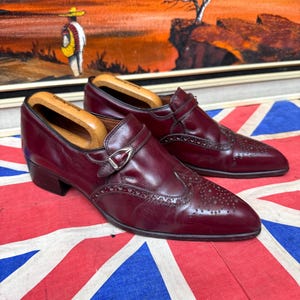 May include: A pair of burgundy leather monk strap shoes featuring brogue detailing on the toe cap and a silver-toned metal buckle closure. Each shoe includes a wooden shoe tree inserted inside the opening to maintain the shape of the footwear.