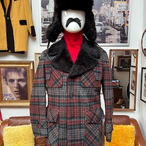 May include: A vintage plaid jacket in red, green, and grey, with a black fur collar and four front pockets. The jacket is displayed on a mannequin wearing a red turtleneck and a fur hat with ear flaps. A yellow jacket hangs in the background.
