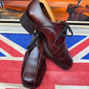 May include: A pair of dark burgundy leather oxford shoes featuring a square toe, a low block heel, and black laces. The shoes are equipped with wooden shoe trees inside to maintain their shape, showcasing a smooth finish and visible stitching.
