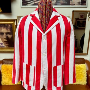 Op de afbeelding: Een rood-wit gestreepte blazer met twee voorzakken en twee knopen. Een paisley sjaal is over de etalagepop gedrapeerd. De blazer wordt gepresenteerd op een etalagepop met een bruine leren bank op de achtergrond.
