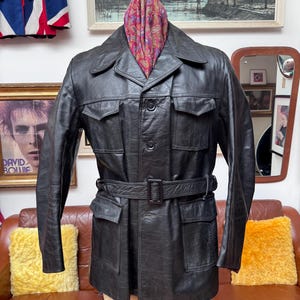 May include: Black leather jacket with a belted waist and multiple pockets. The jacket has a classic design with a collared neckline and button-down closure. A colorful paisley scarf is draped around the neck. The jacket is displayed on a mannequin.