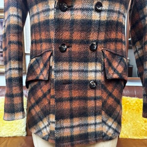 May include: A double-breasted plaid coat in shades of brown, orange, and black. The coat features two rows of black buttons, two front pockets, and a classic collar. The fabric has a textured appearance.