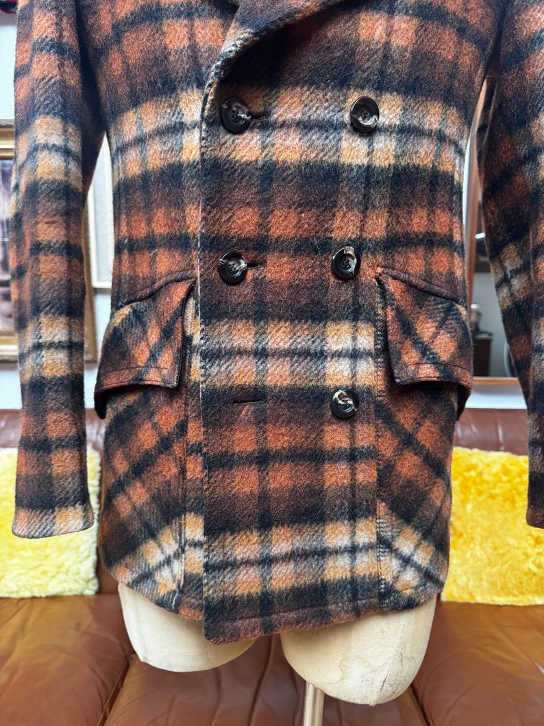 May include: A double-breasted plaid coat in shades of brown, orange, and black. The coat features two rows of black buttons, two front pockets, and a classic collar. The fabric has a textured appearance.