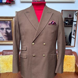 May include: A brown double-breasted blazer with gold buttons, a red turtleneck, and a patterned pocket square. The blazer is displayed on a mannequin. The background includes framed artwork and a Union Jack flag.